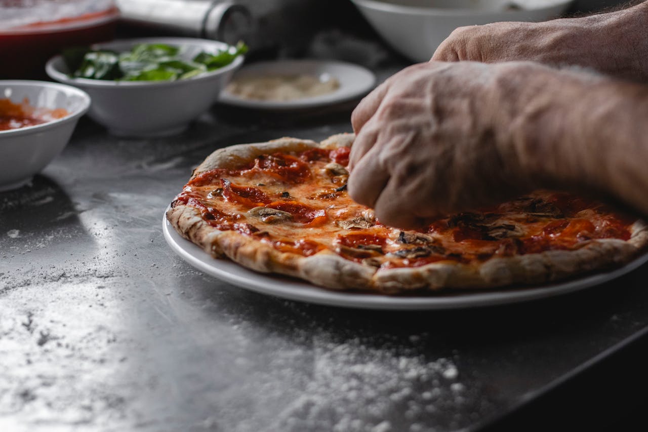 Close-up of man's hands preparing a freshly cooked pizza with toppings in the kitchen.